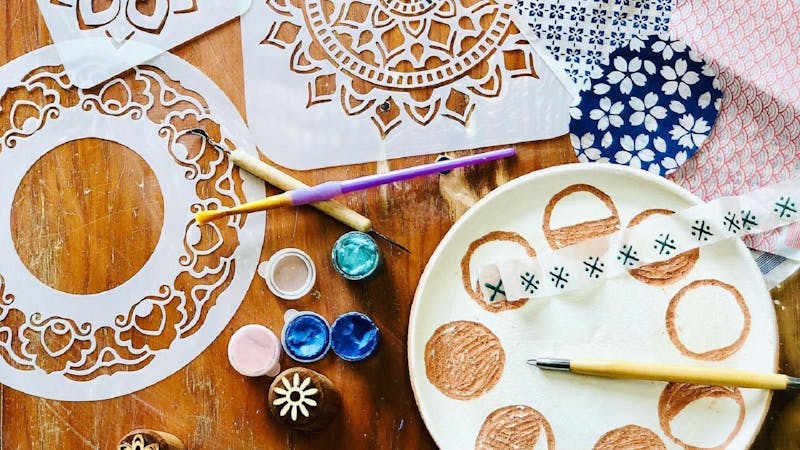 Stencils, underglazes, tools and a ceramic plate are shown in flatlay form