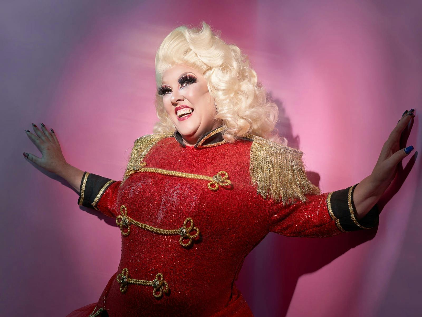 Drag Queen in Red Jacket standing against a pink wall smiling with a blonde wig
