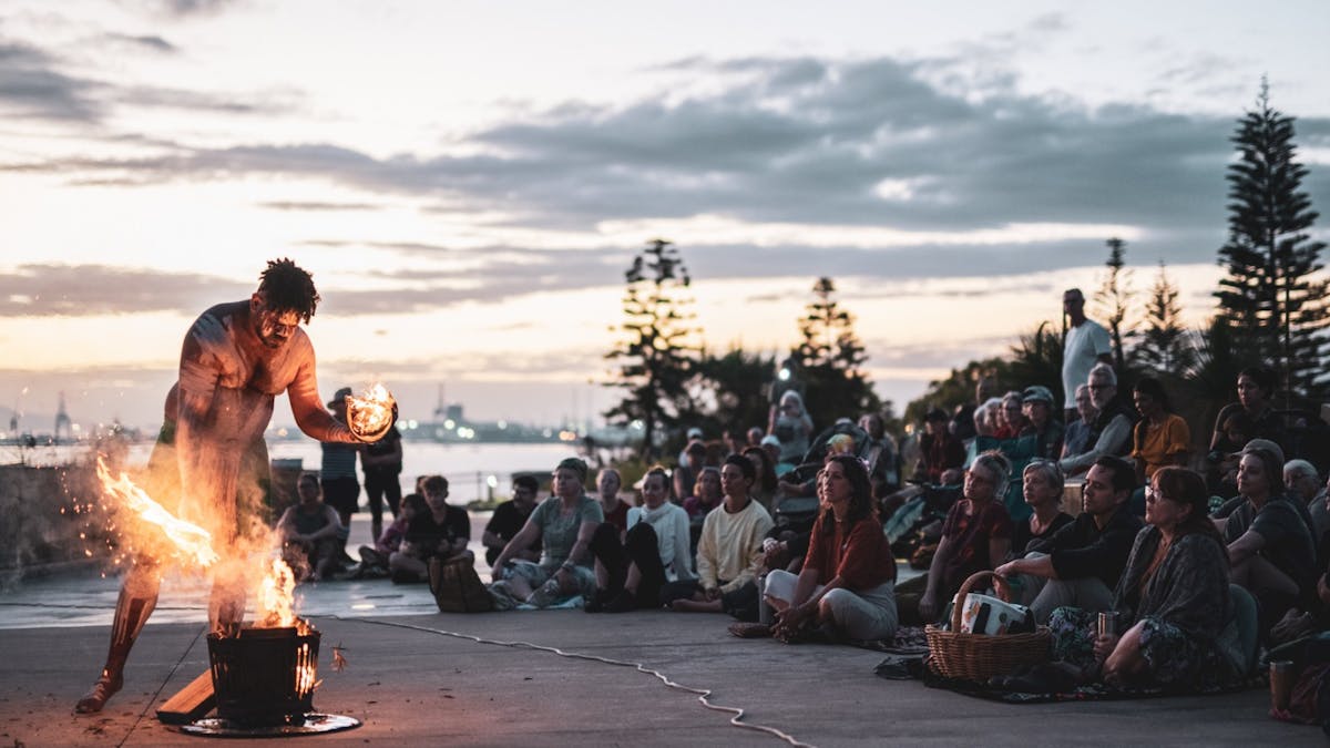 A First Nations performer lights a fire as a crowd watches on at sunrise.