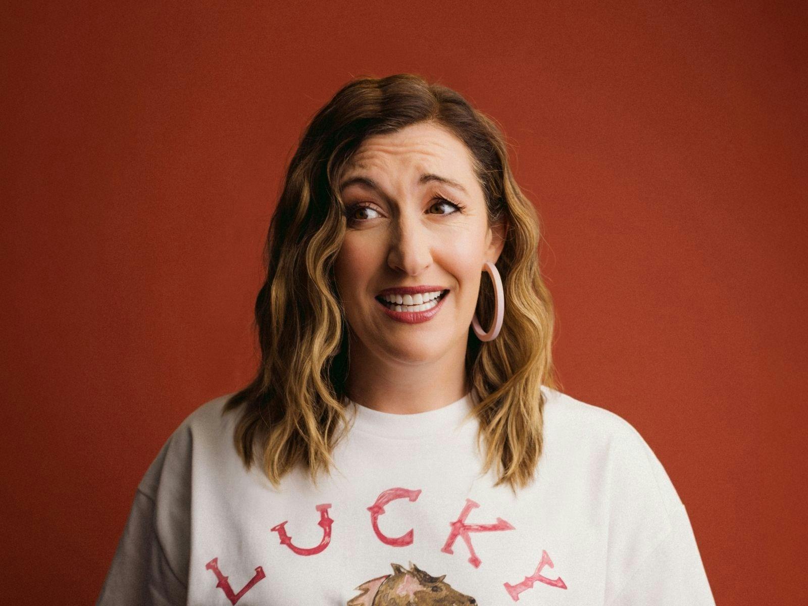 Celia Pacquola smiling against a warm red backdrop, wearing a white T-shirt and hoop earrings.