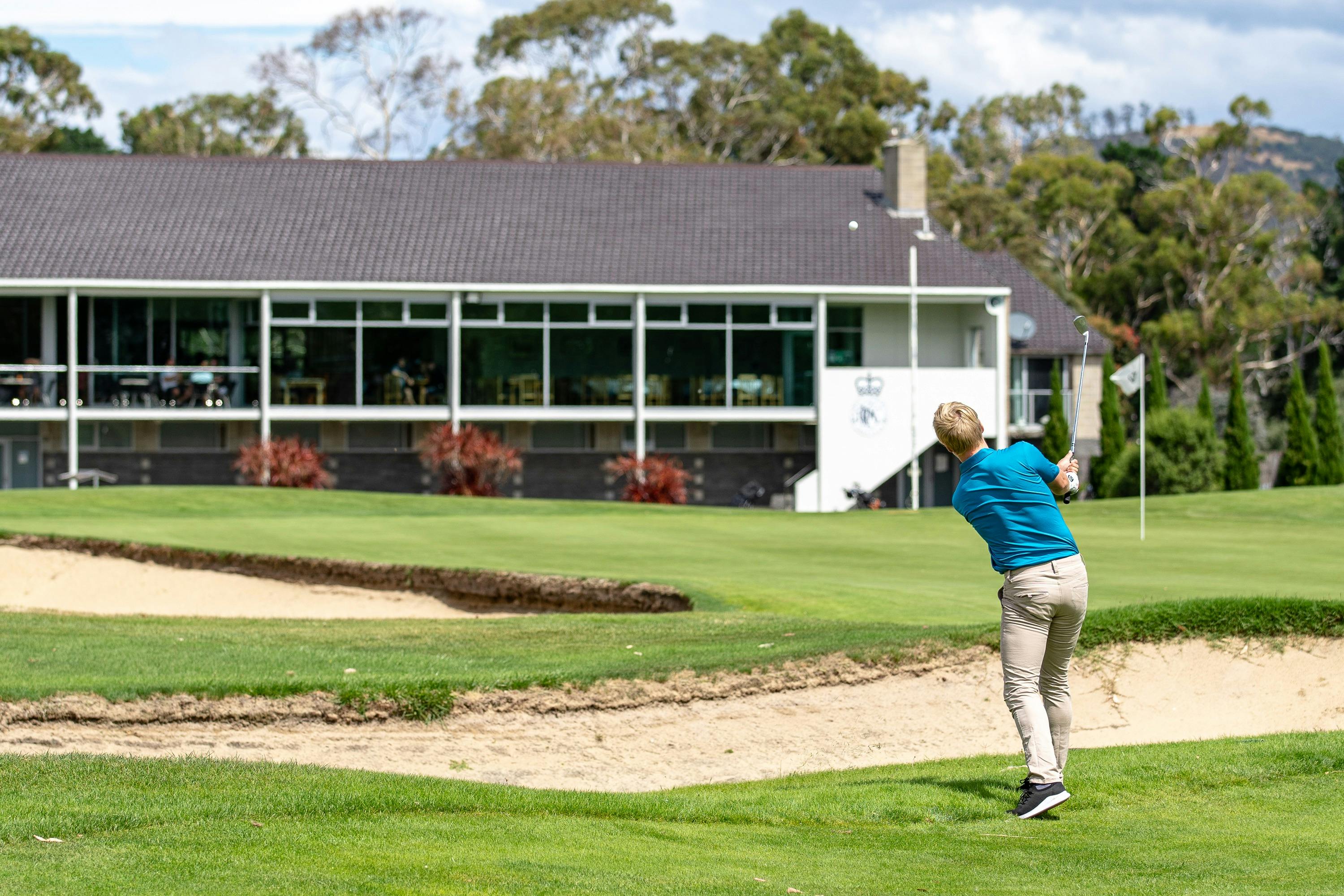 Golfer hitting ball over greenside bunkers, with clubhouse in background