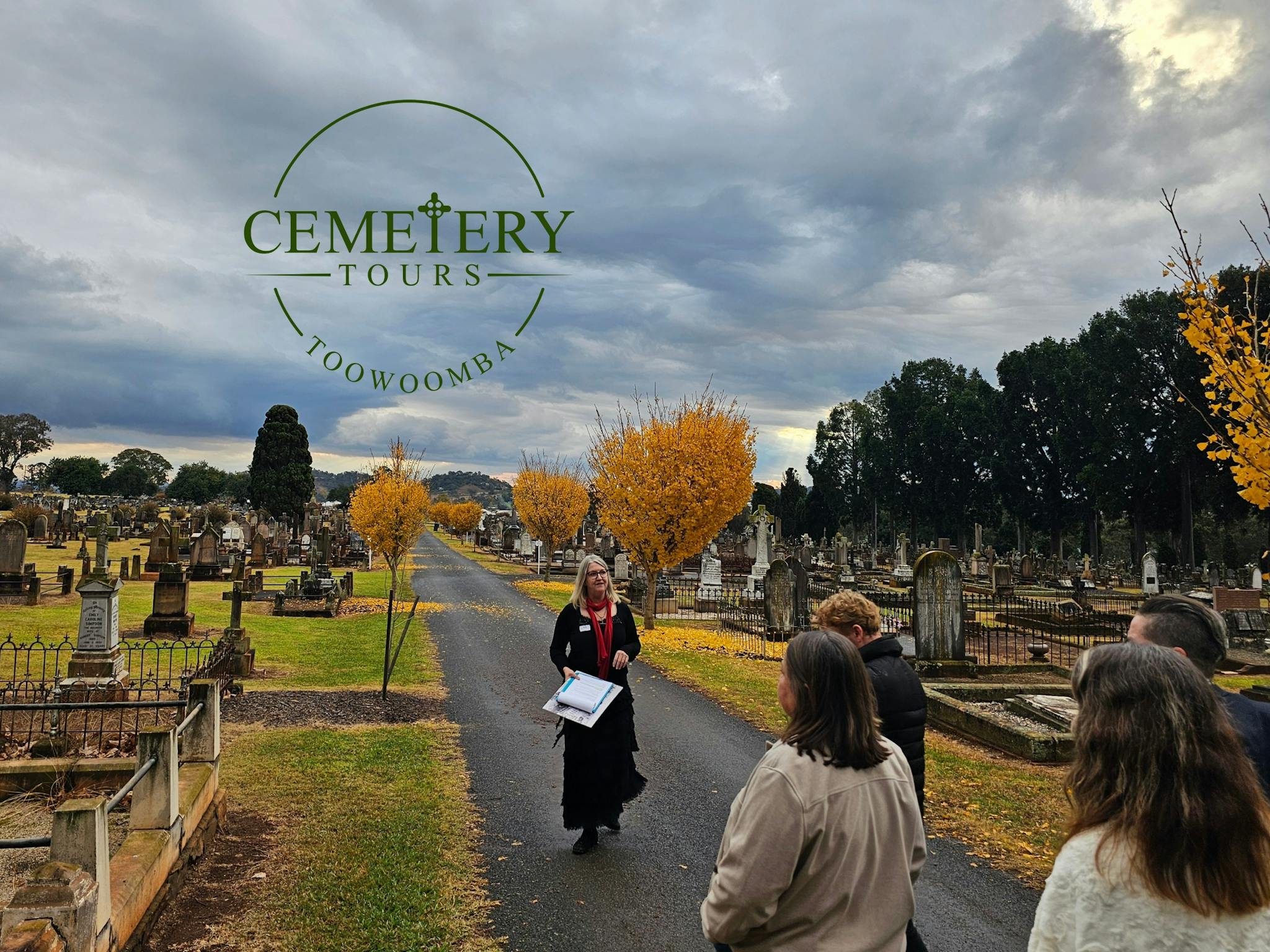 A guide stands in the cemetery,  trees with bronze autumn leaves, people listening to guide