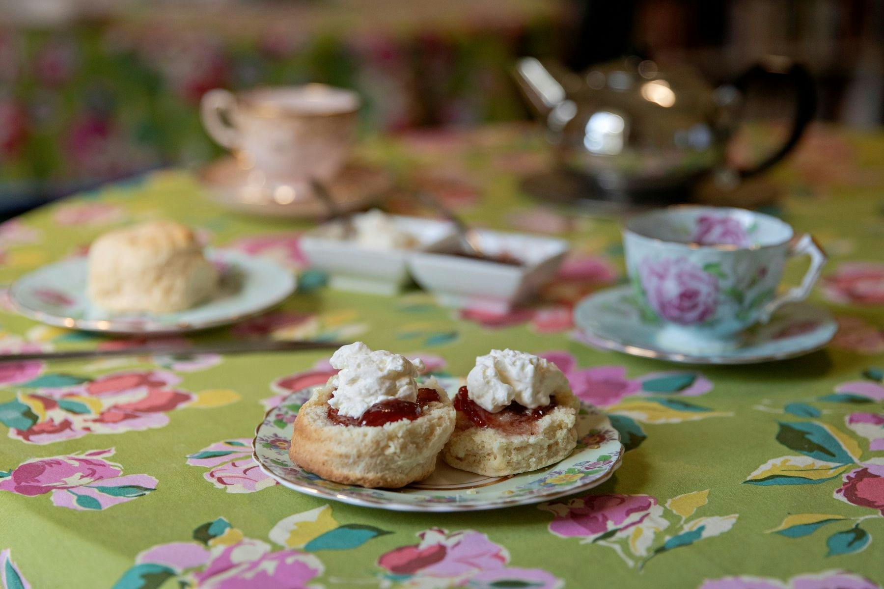 A pair of scones with jam and cream in front. Tea cups and pot with more nibblies just behind.