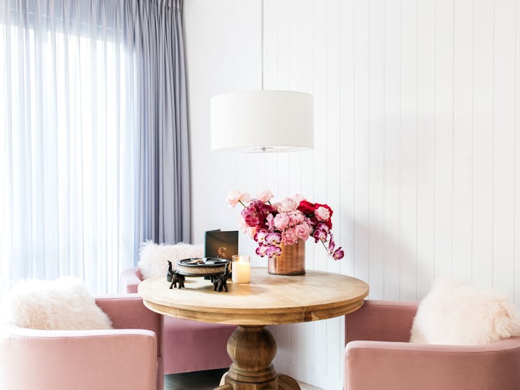3 pink chairs surround a wooden table with fluffy cushions on them and pink flowers on the table
