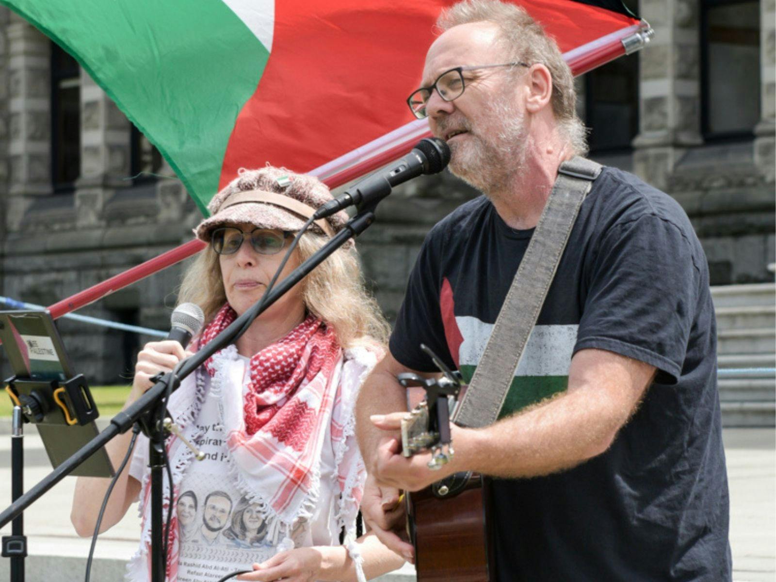 David Rovis & Kamala Emanuel singing live at a protest in front of a Palestinian flag.]
