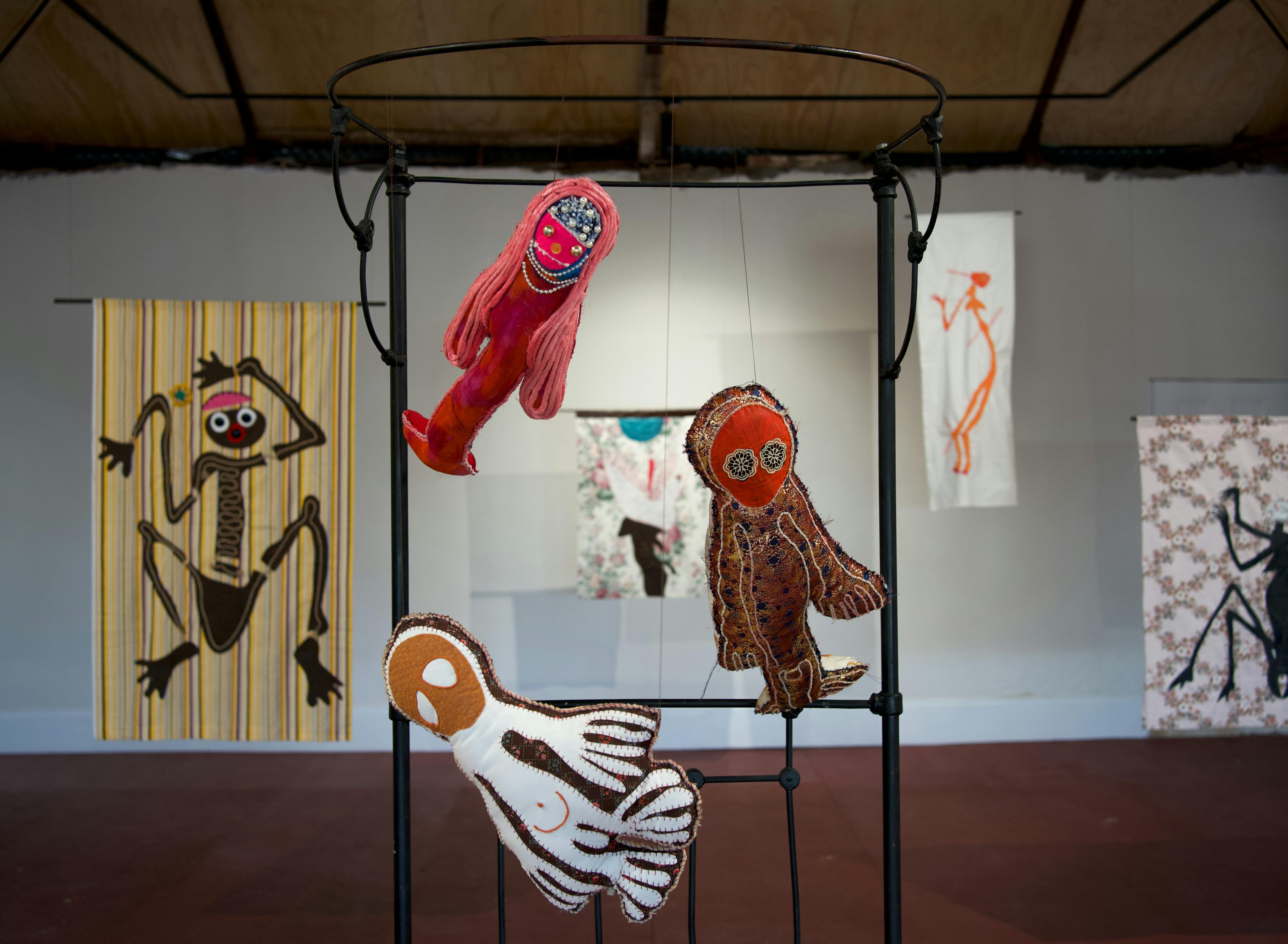 A bedframe suspends 3 soft sculptures, textile works hang in the background in a white wall gallery
