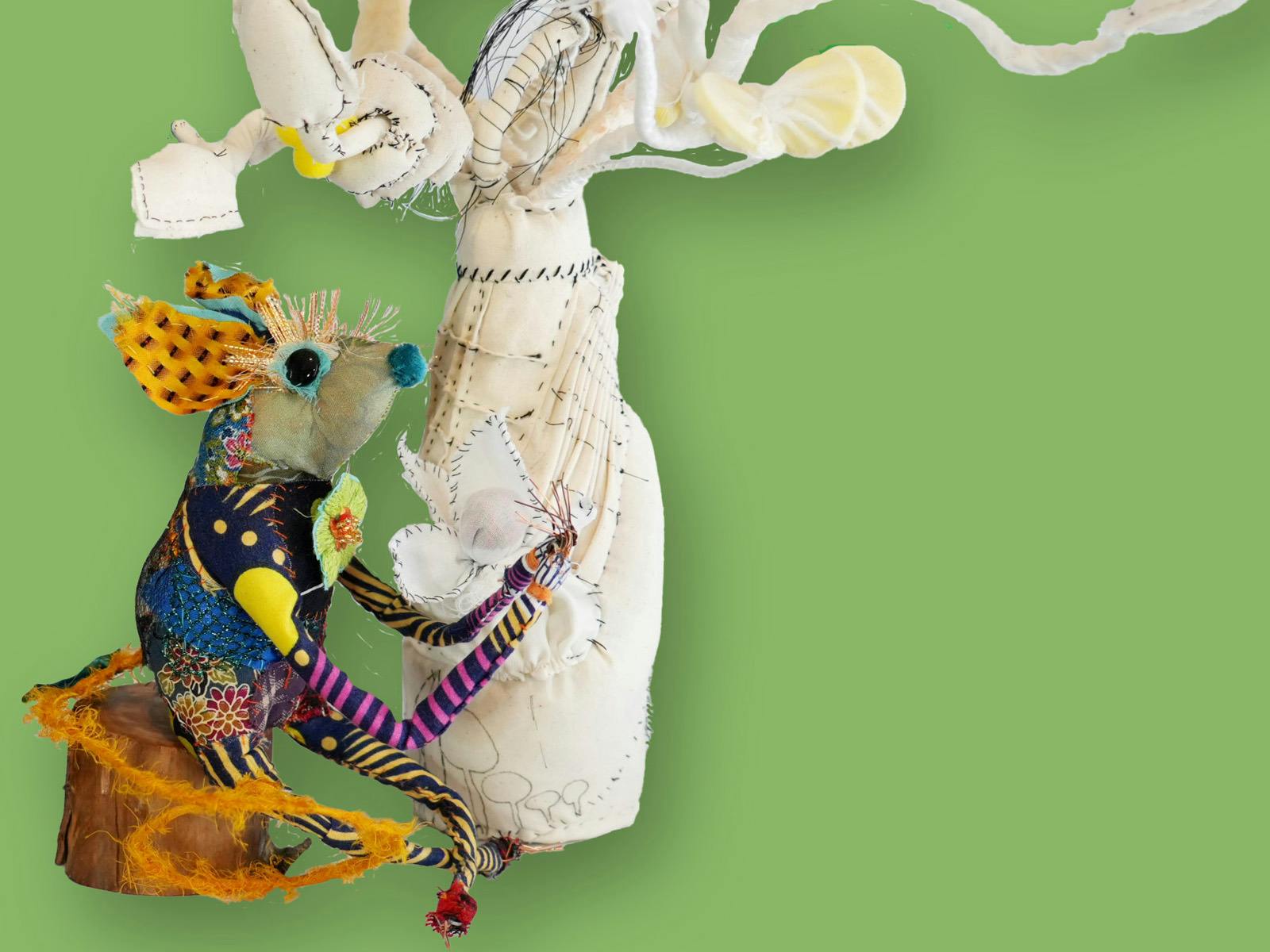 A colourful textile mouse figure sits in front of a twisted white plant sculpture.