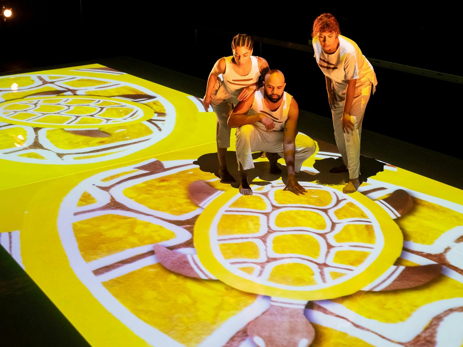 Lit by a projection from above, three people look down at a yellow projection of a turtle.