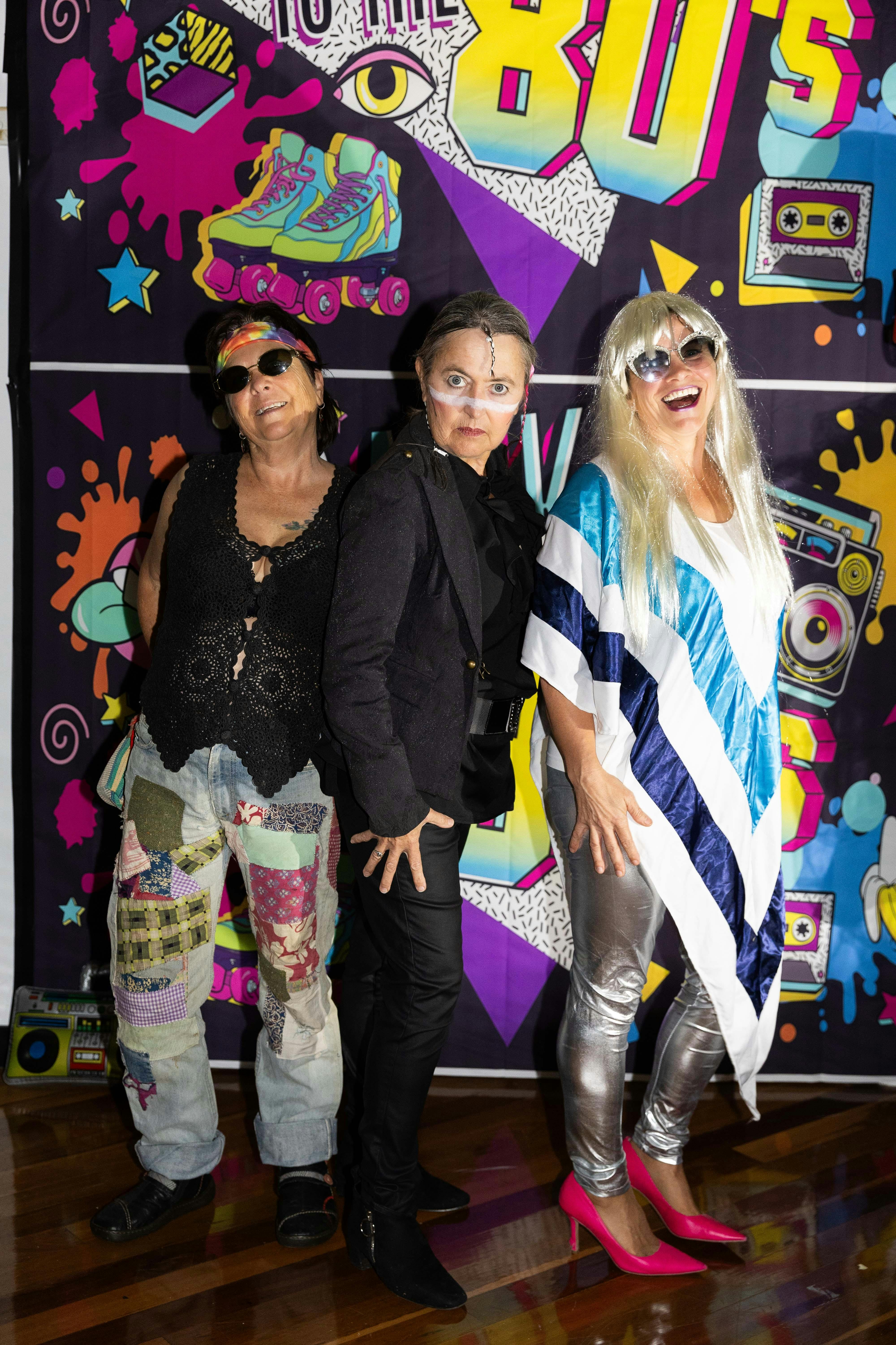 Three ladies pose in front of an 80s backdrop