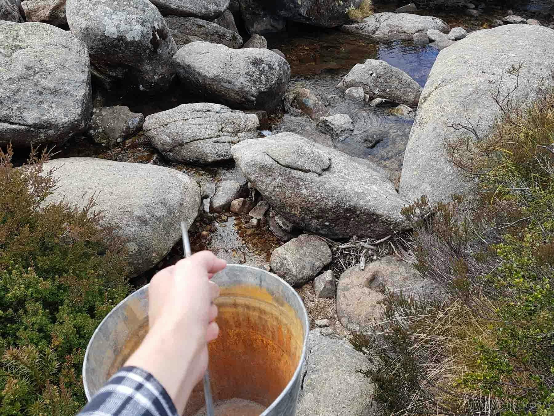 A first person view - hand carrying an empty metal bucket and approaching a rocky stream.