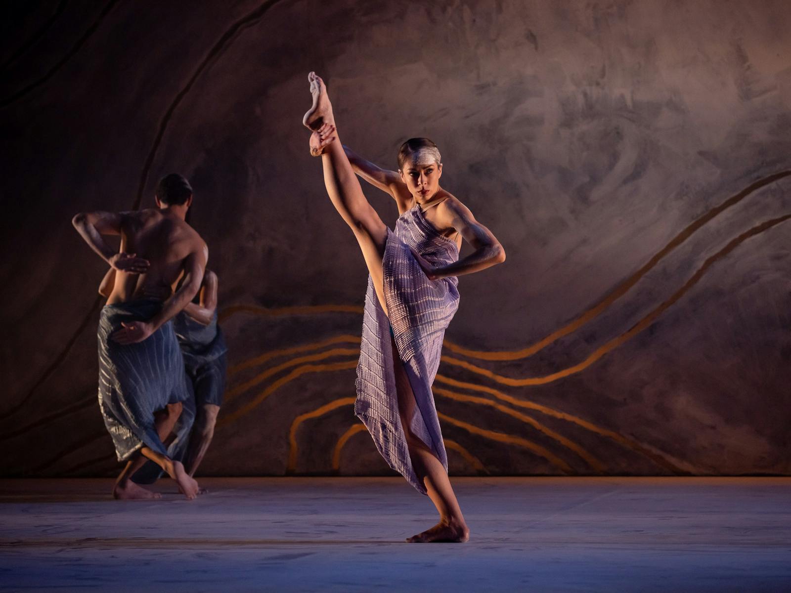 A dancer in a purple dress lifting her leg on stage with two dancers in the background