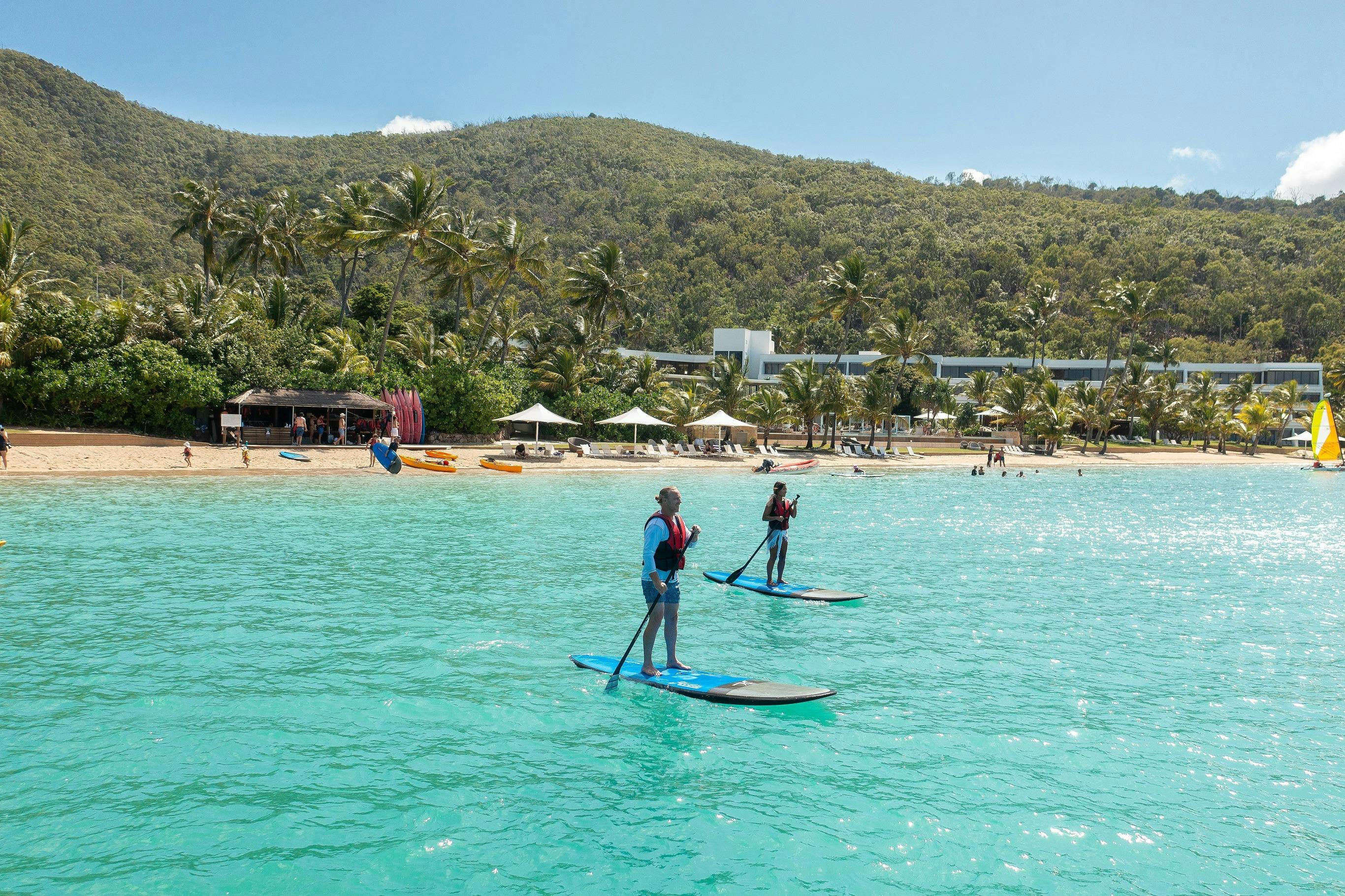 Two people paddleboarding in the ocean waters - a resort beach and hills in background