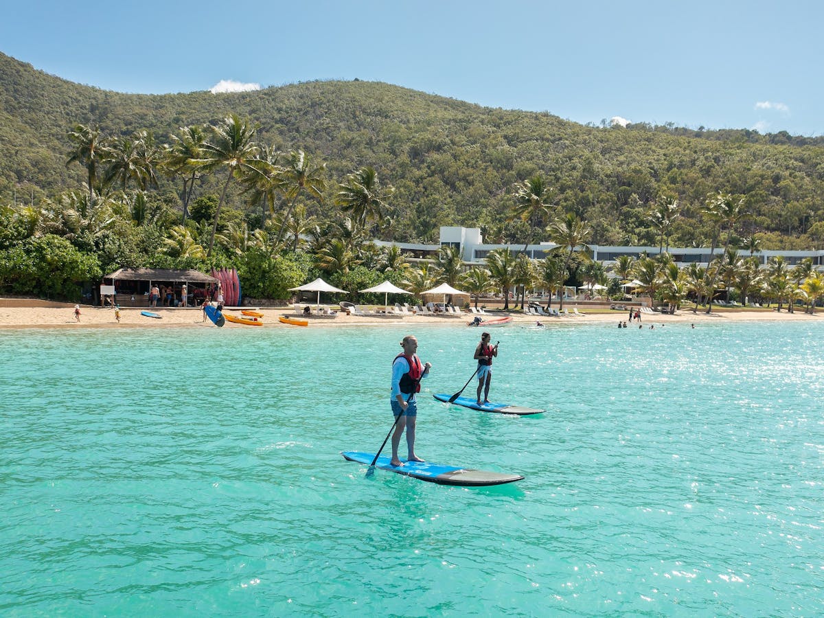 Two people paddleboarding in the ocean waters - a resort beach and hills in background