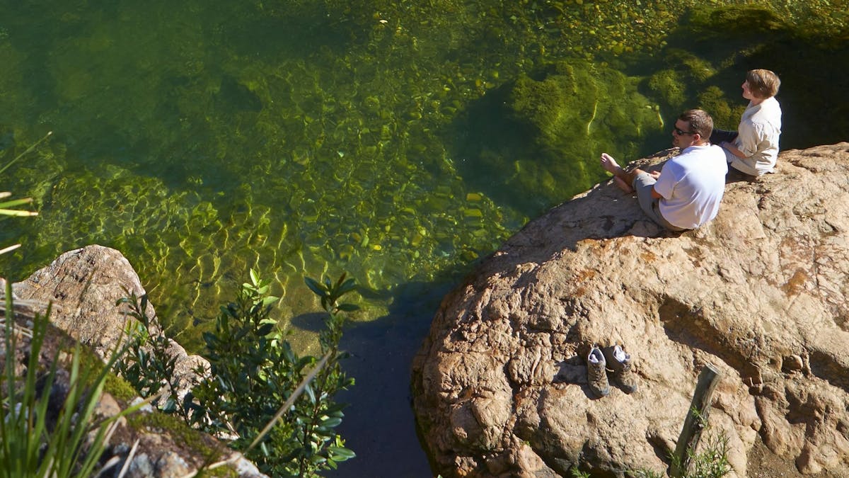 Two people sitting on a rock with their bare feet dangling over into a waterhole.