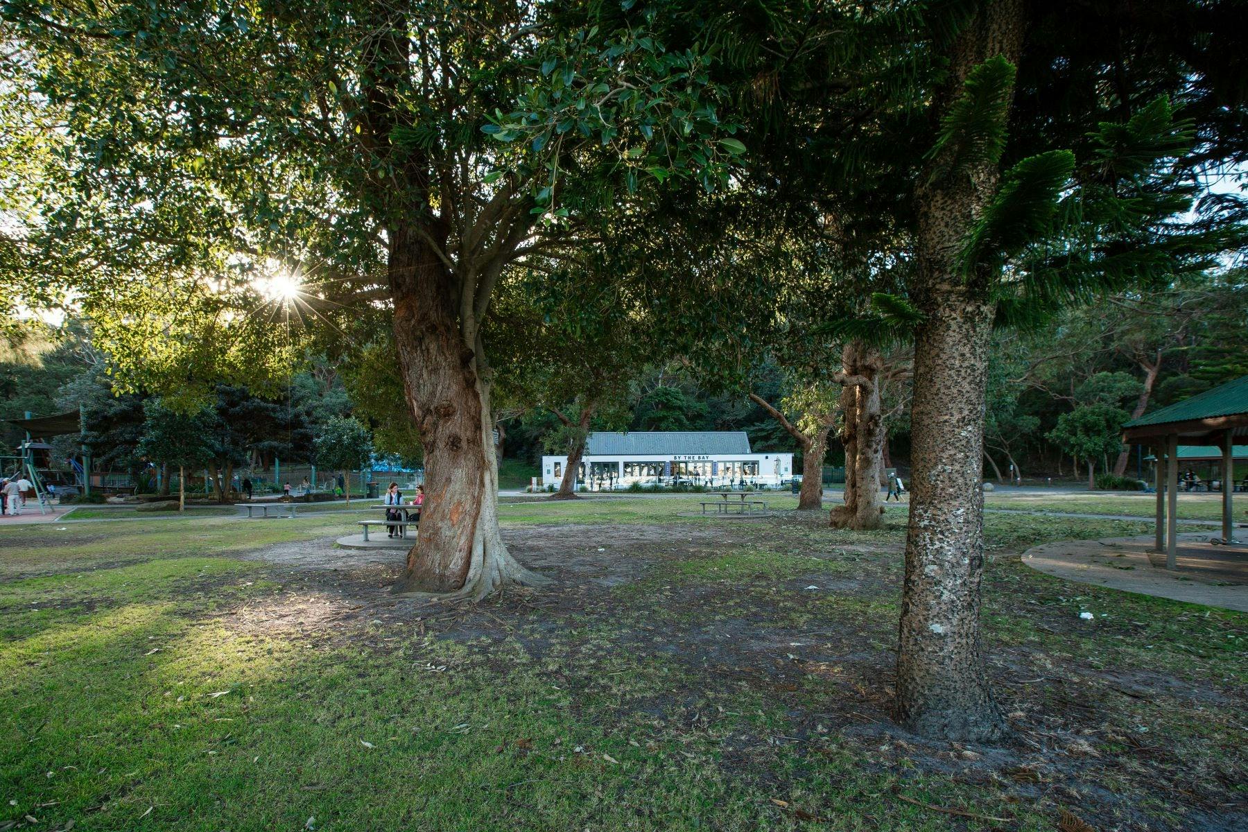 A shaded area with trees, a bench with people sitting and a cafe building  behind them.