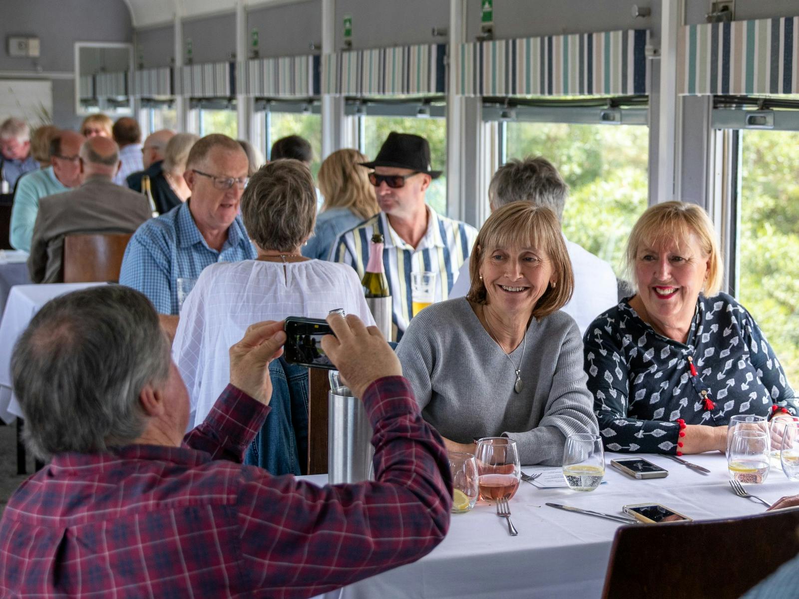 Inside a train carriage a man takes a photo of two women at a table