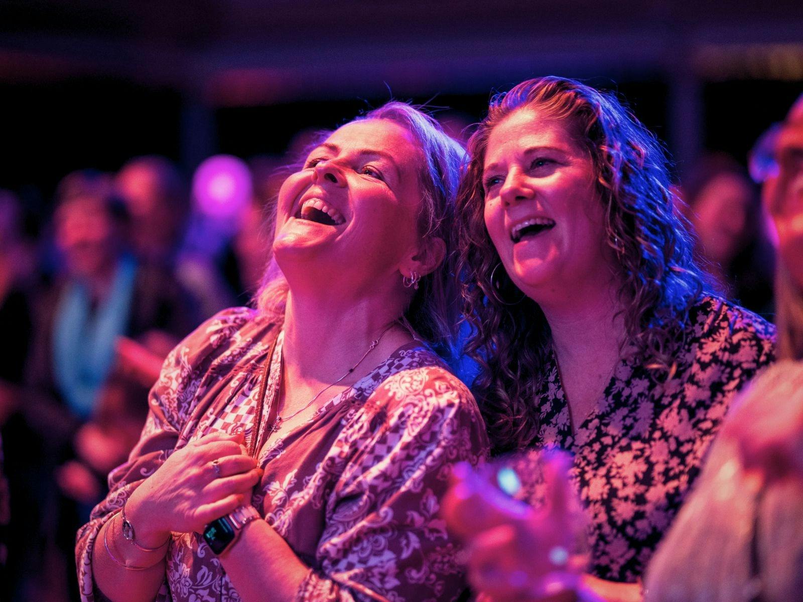 Two women laughing and smiling brightly in the audience under colorful stage lights
