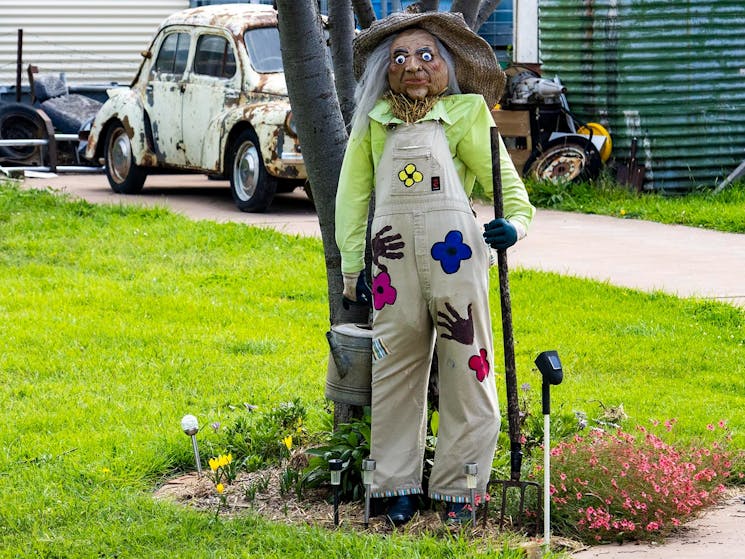 a scarecros dressed in overalls standing in front of a tree holding a watering can