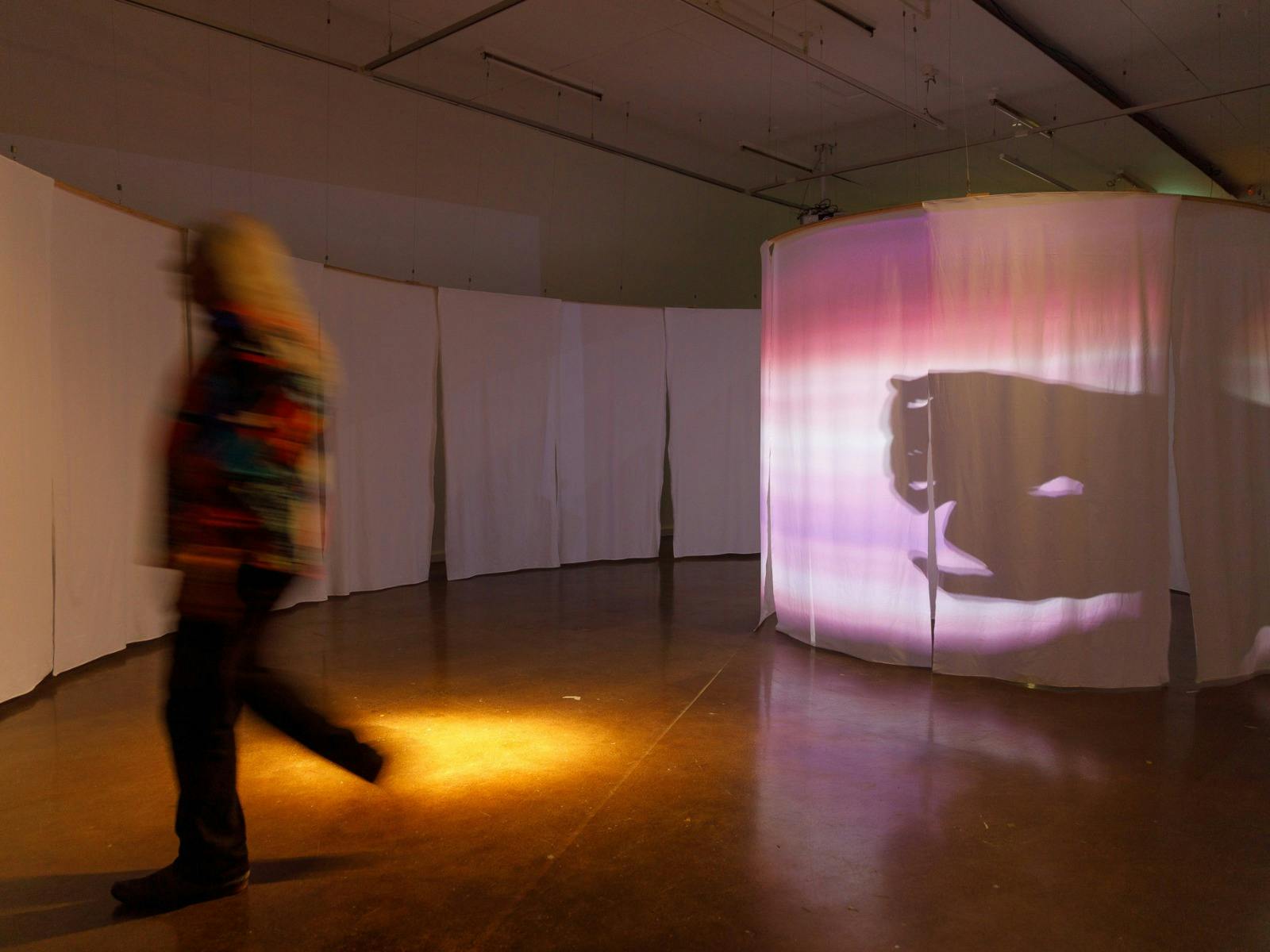 A visitor walks through a dimly lit exhibition space featuring flowing white screens and projected i