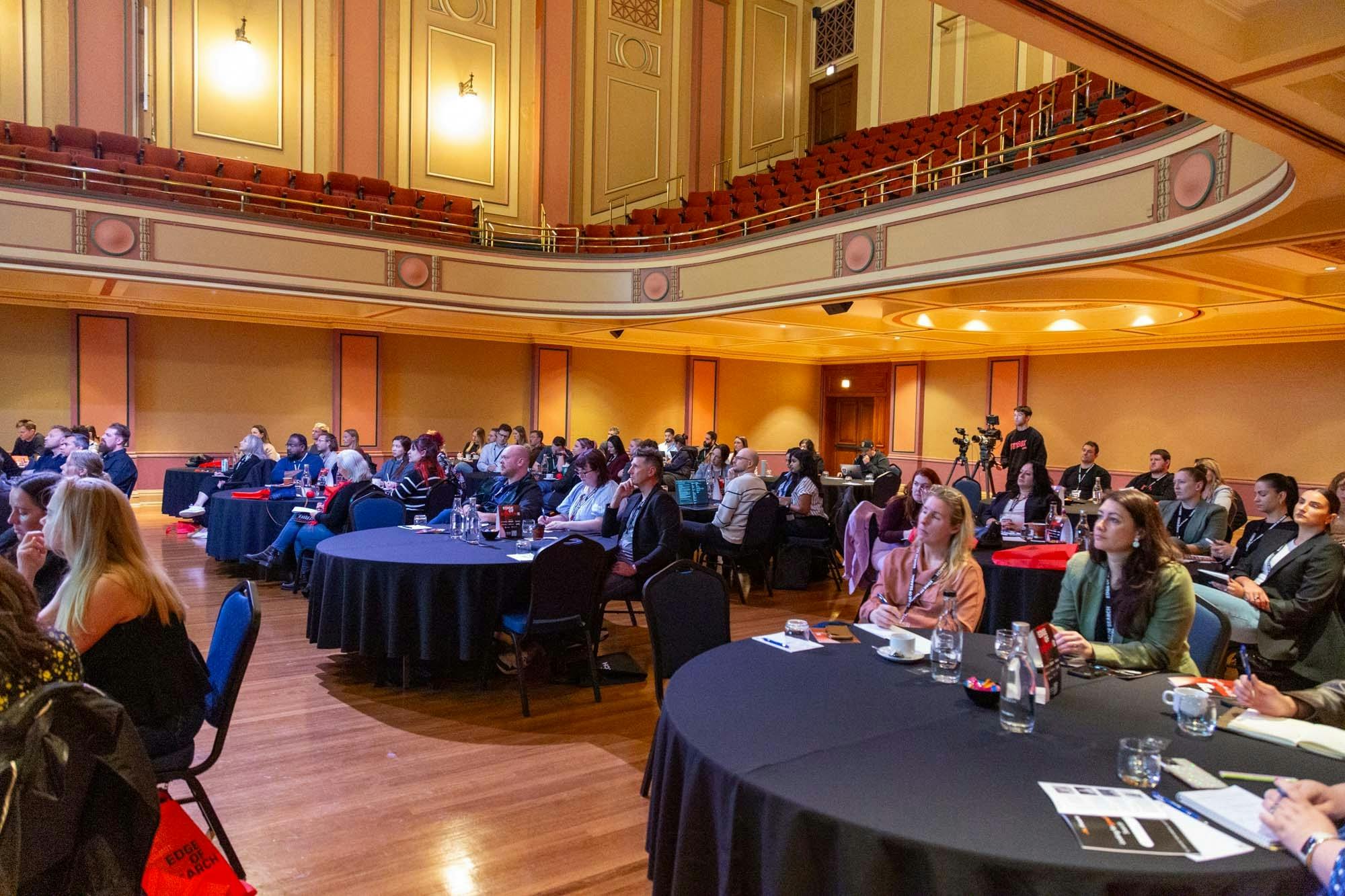 People in a large hall with balcony seating, sitting at round tables covered in black tablecloths.