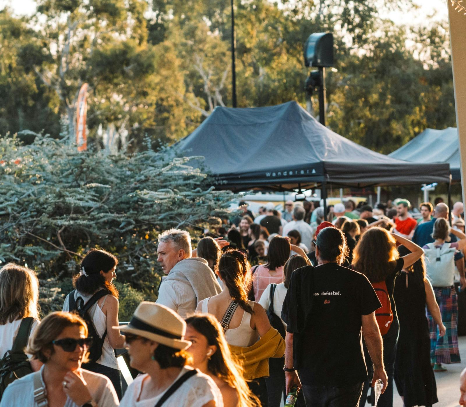 A bustling crowd outdoors at sunset. Tents and trees are visible,