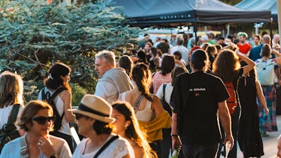 A bustling crowd outdoors at sunset. Tents and trees are visible,