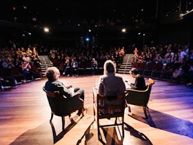 Two individuals seated on chairs, addressing an audience in a formal setting.