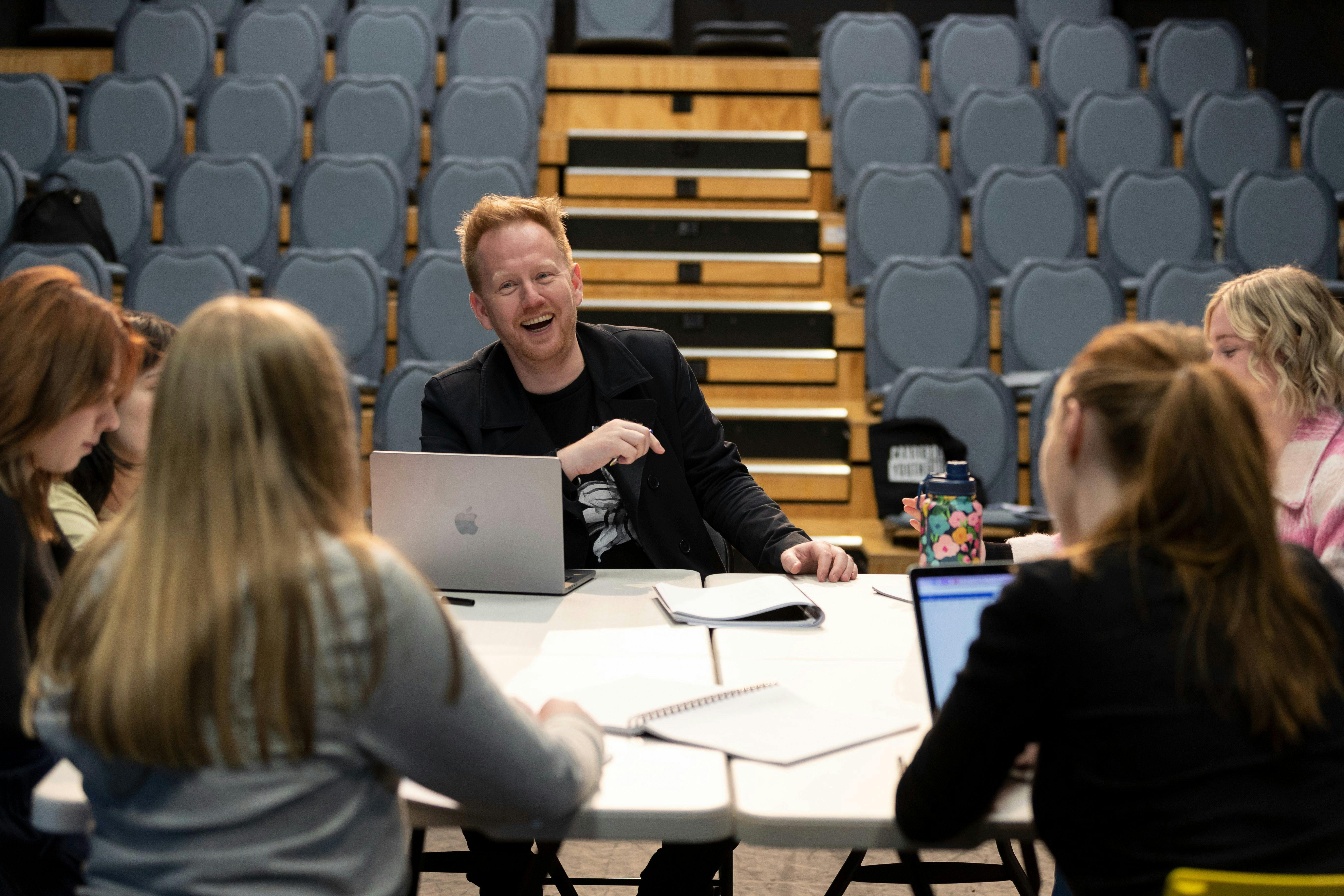 A man, laughing, sits at a table with a group of teenagers with laptops and notebooks in a theatre.