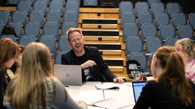 A man, laughing, sits at a table with a group of teenagers with laptops and notebooks in a theatre.