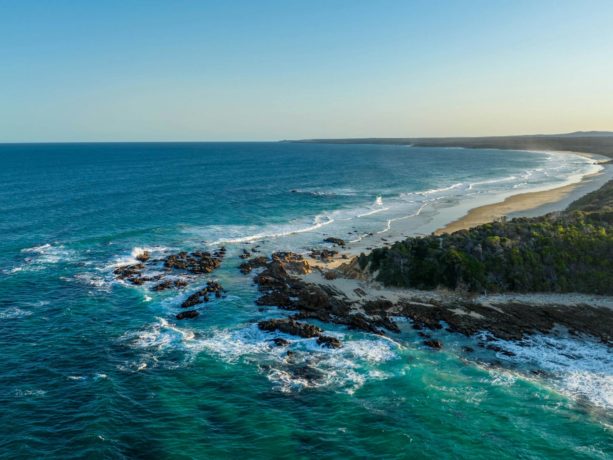 Aerial view of a curving coastline with lush greenery, sandy beach, and rocky areas, waves crashing
