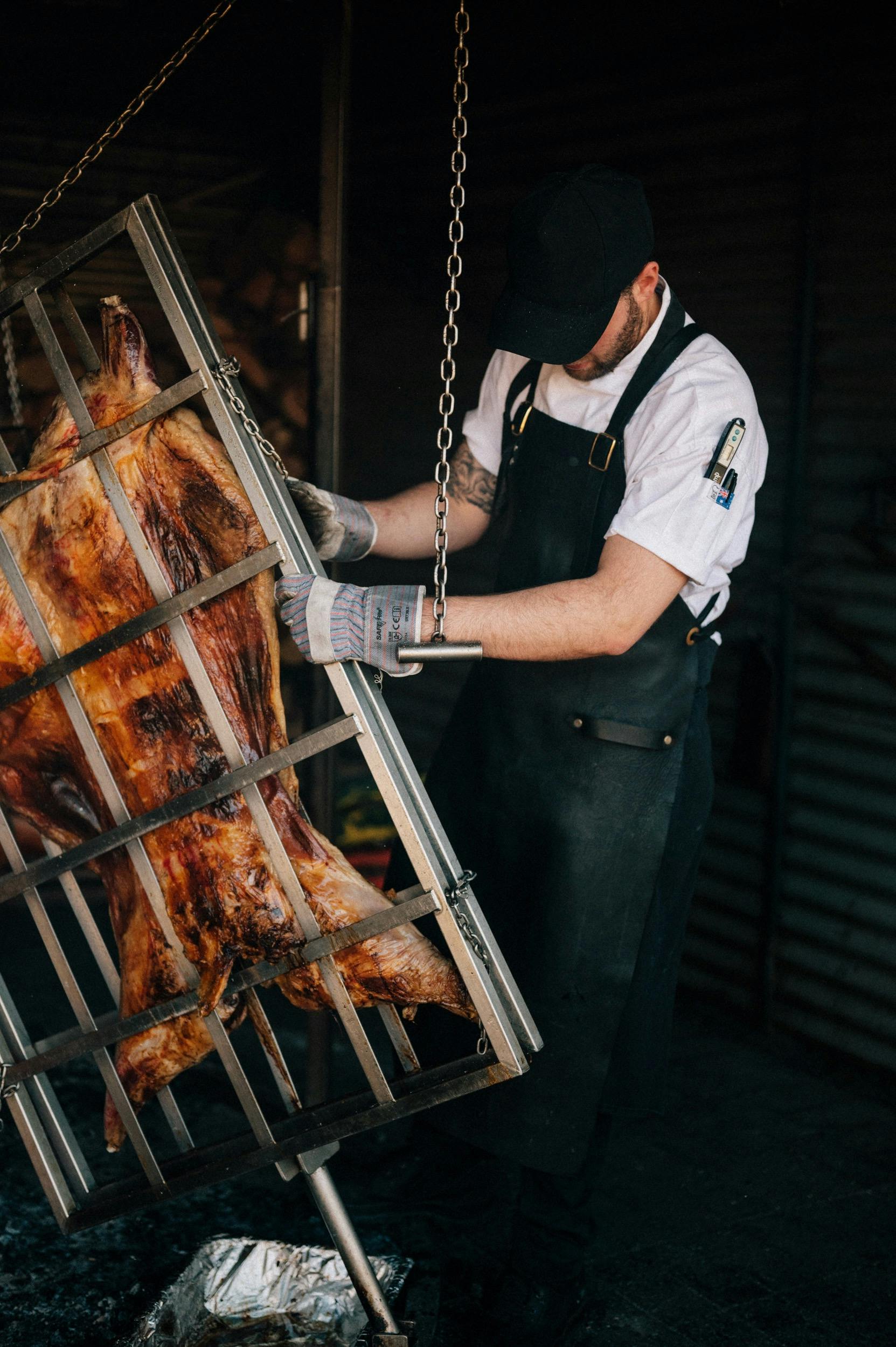 A chef manoeuvres a metal frame cooking meat over coals