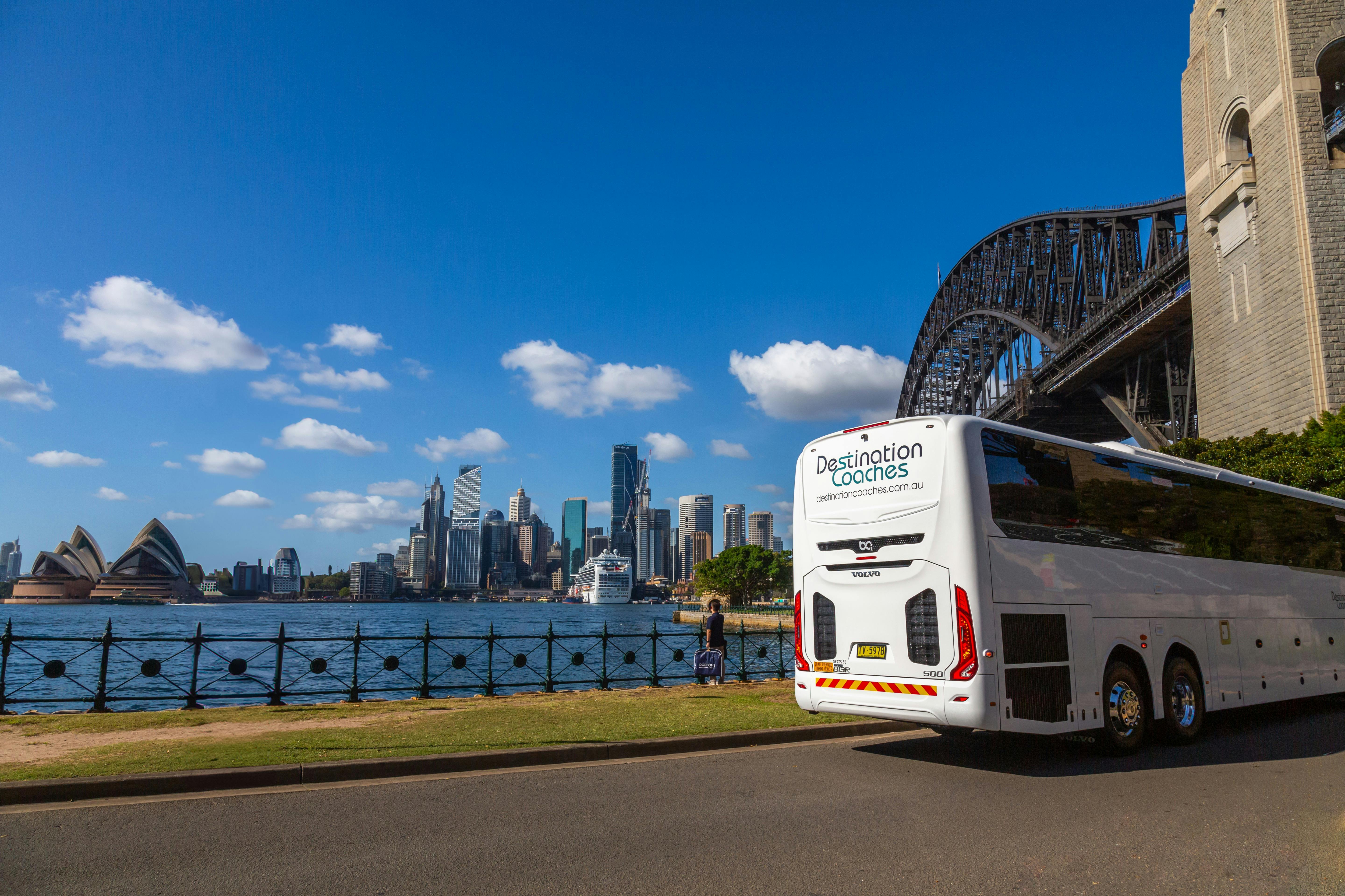 Reisebus neben ruhigem Hafenwasser mit Brücke und Stadtgebäuden.