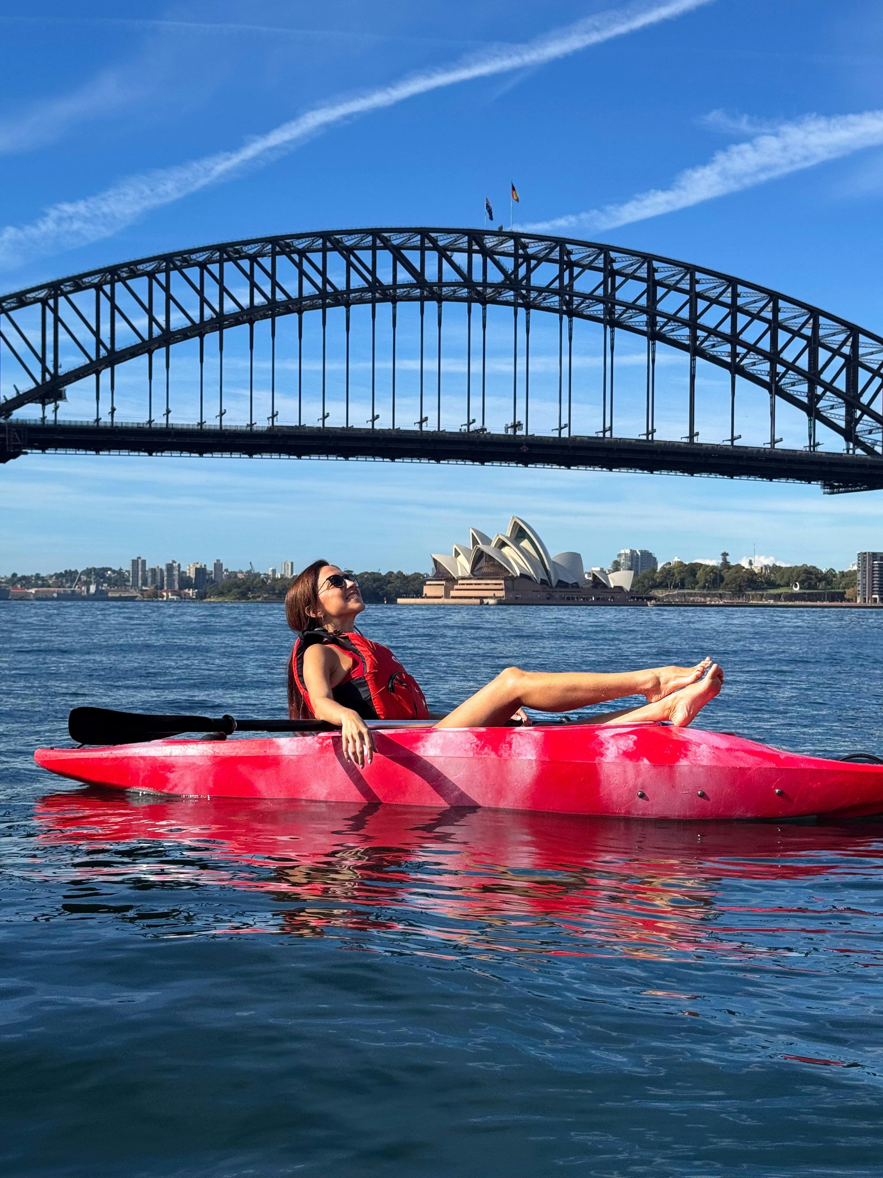 Lady laying back with legs out of kayak sun bathing with Sydney icons in background on a sunny day.