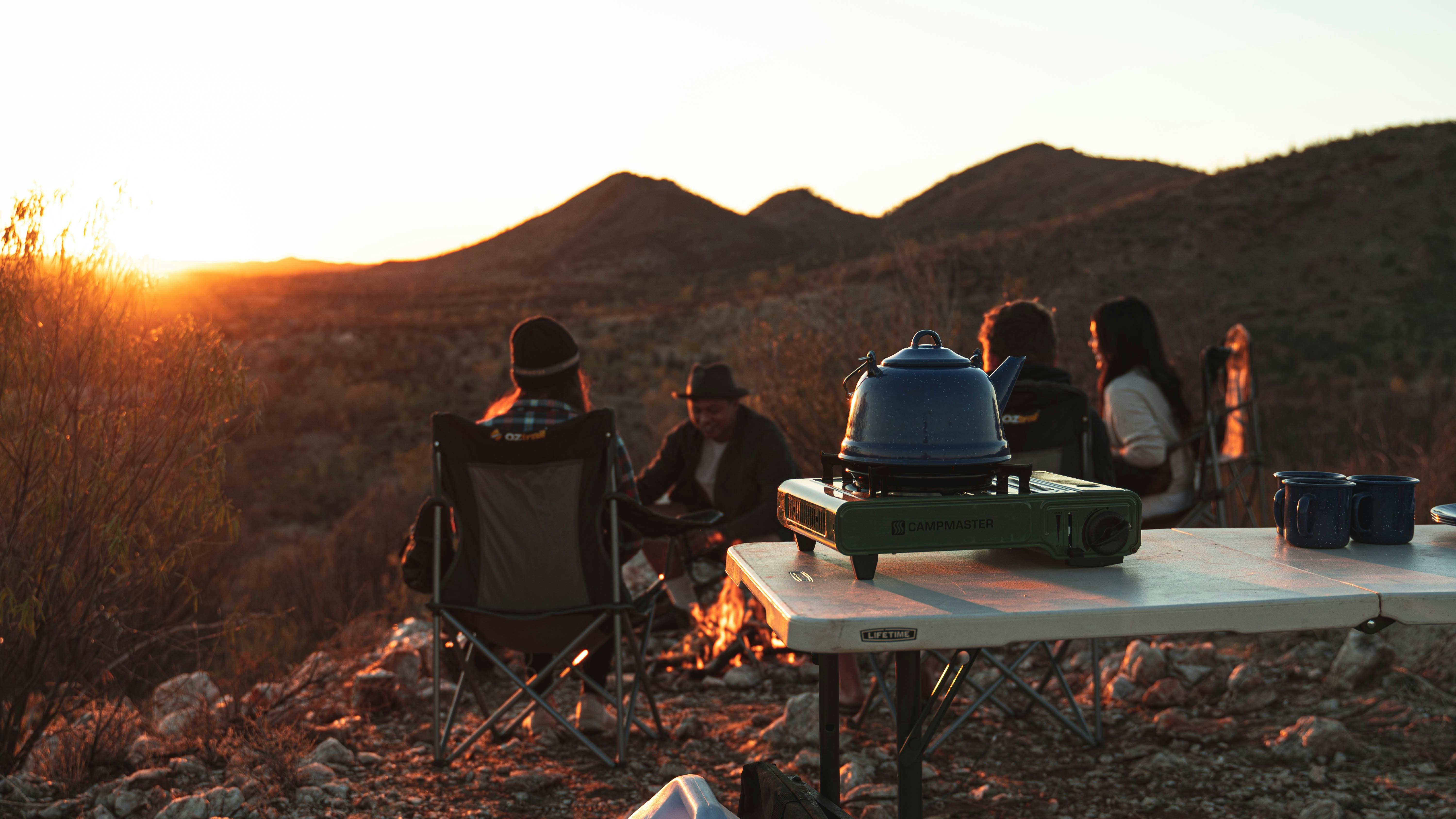 Camping stove and billy with campers in the background sitting around a fire looking at the sunset