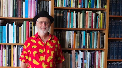 An elderly man wearing a black hat and a banana printed shirt while standing in front of a bookcase