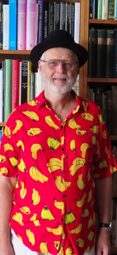 An elderly man wearing a black hat and a banana printed shirt while standing in front of a bookcase