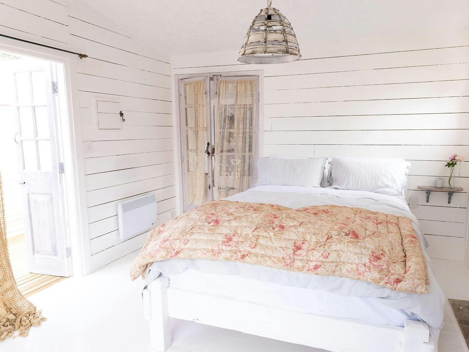 A large bed with white linen and a pink blanket in a simple cosy room with wooden doors & white wall
