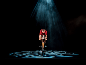 A man lit by a blue textured spotlight is riding a bicycle on stage wearing a red riding suit.