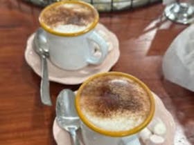 Two delicious mugs of cappuccino on pink saucers with marshmallows and chocolate powder on top.