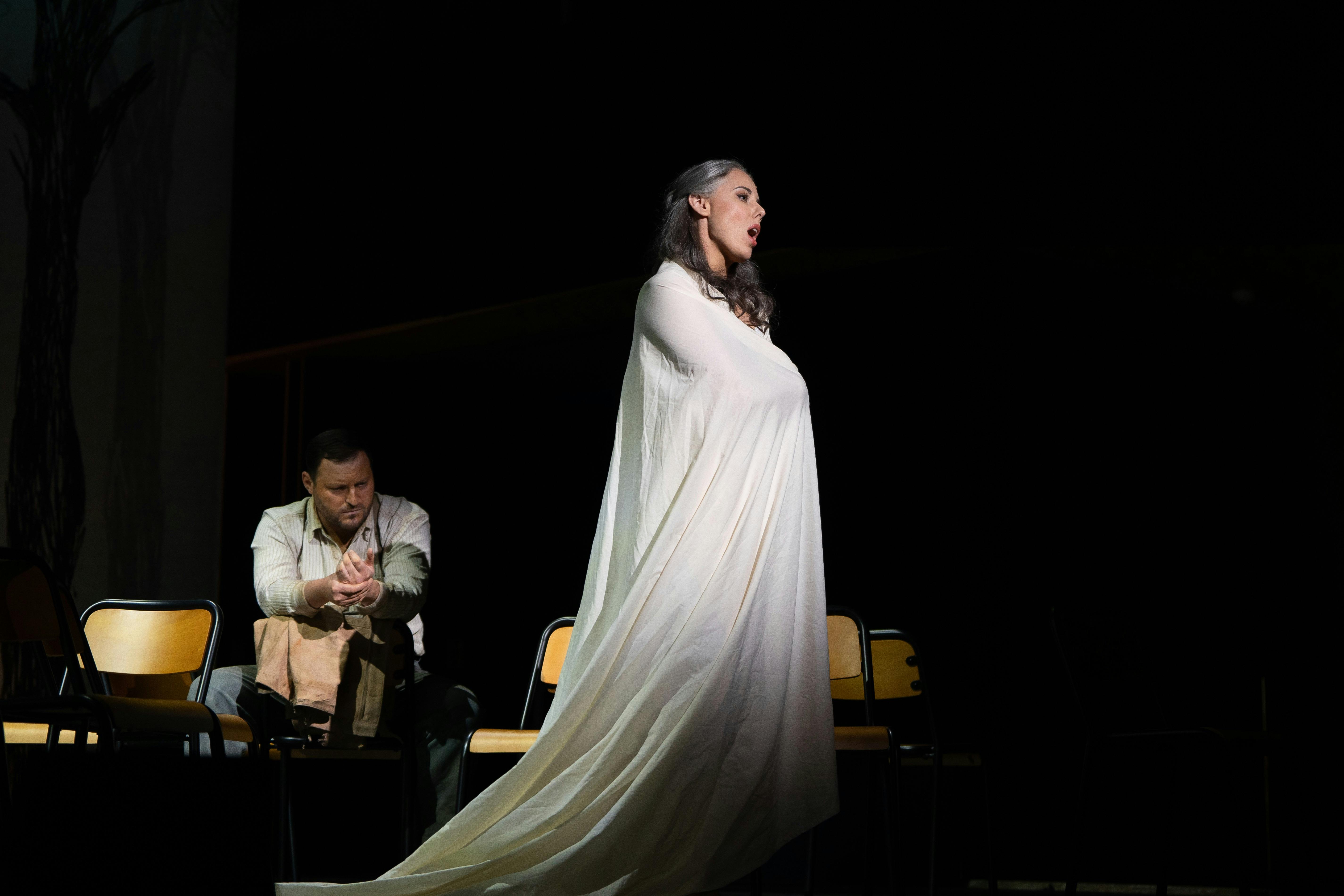 Performer draped in white stands in spotlight as another sits among chairs on stage.
