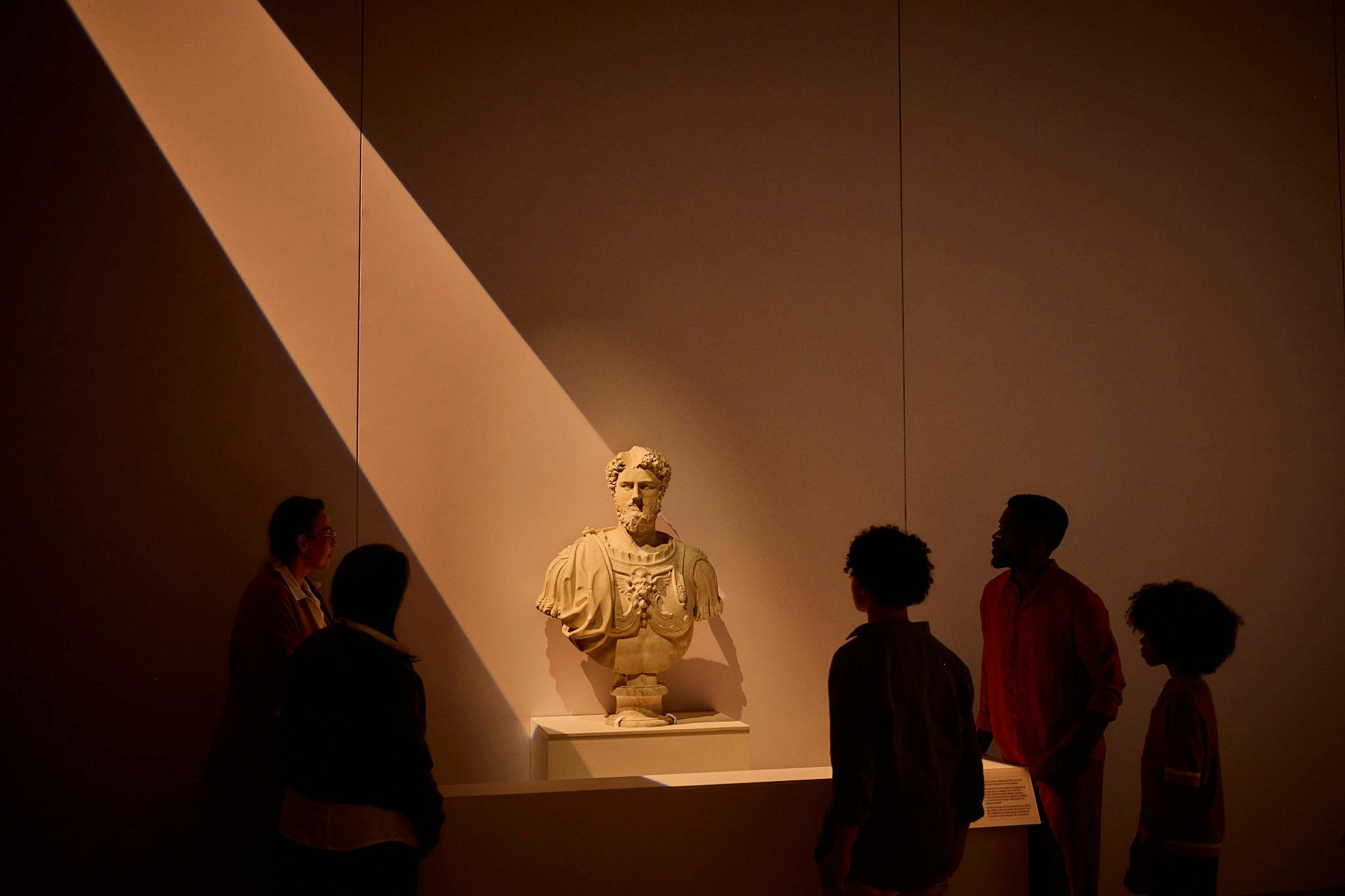 Group of visitors standing around a marble Roman bust on a plinth under dramatic overhead lighting.