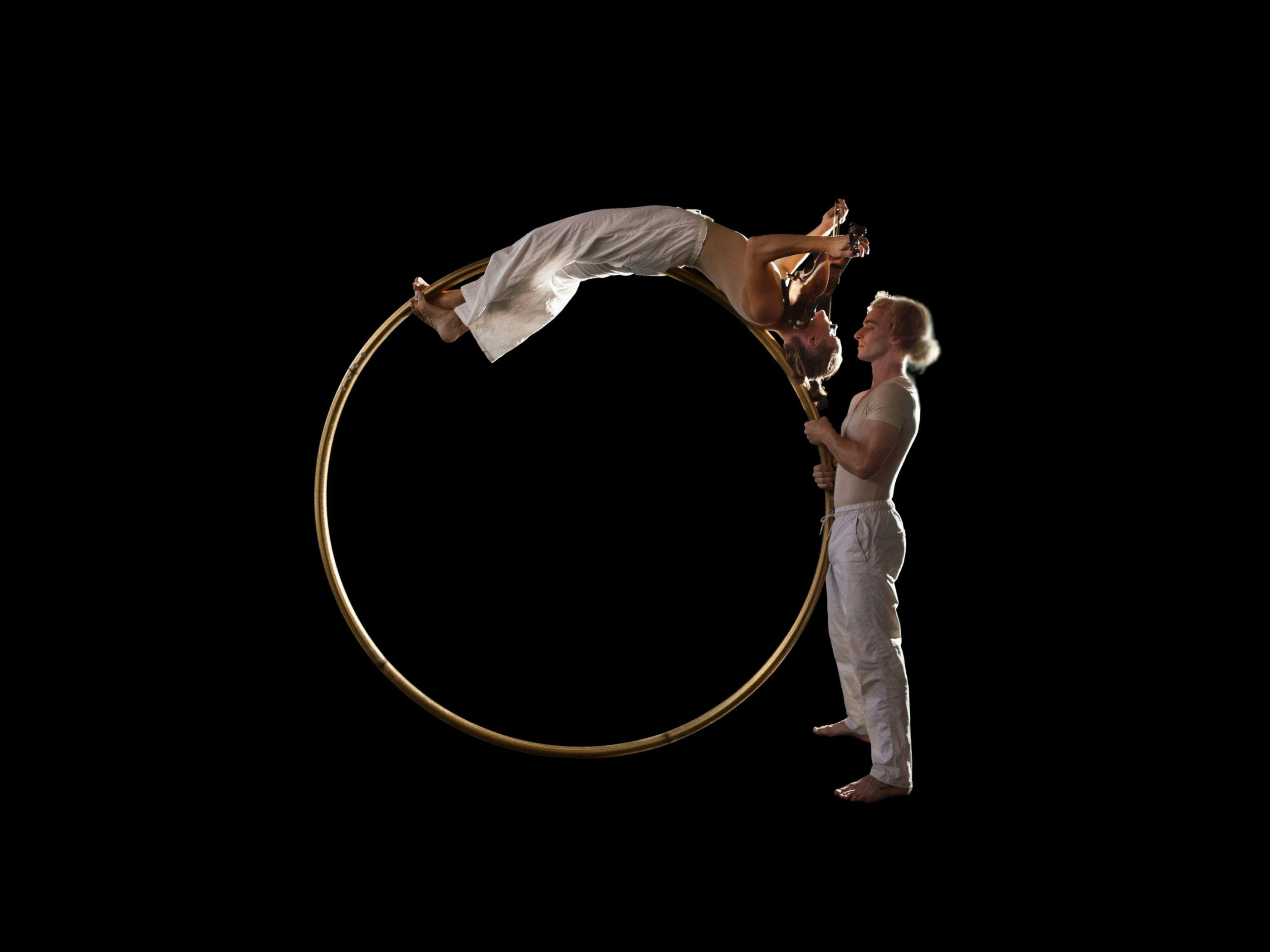 An acrobat holds a circus hoop upright, while an acrobatic violinist balances on top with her violin