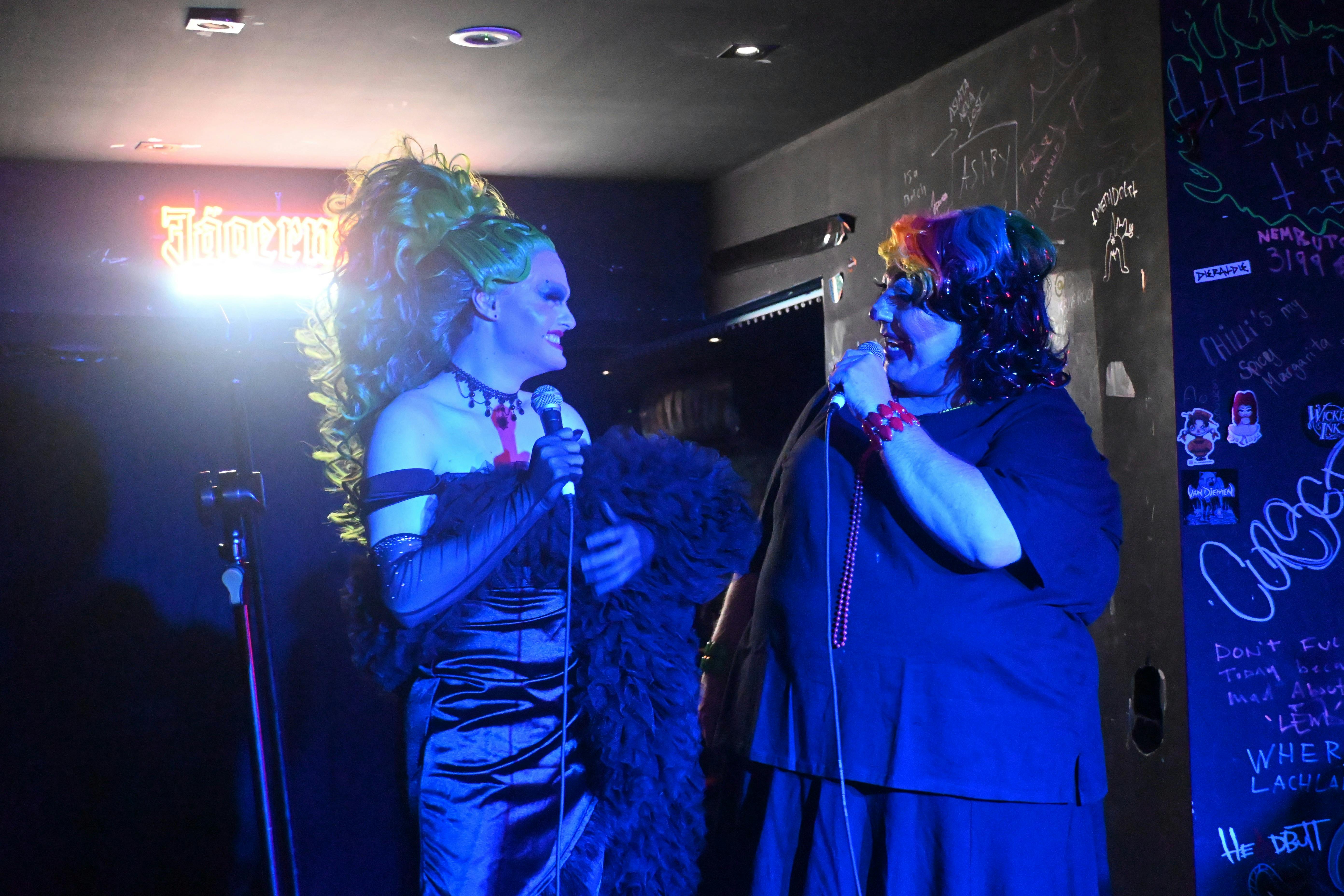 Two drag queens, a slim one and a plus-size one, converse on stage using microphones and smiling
