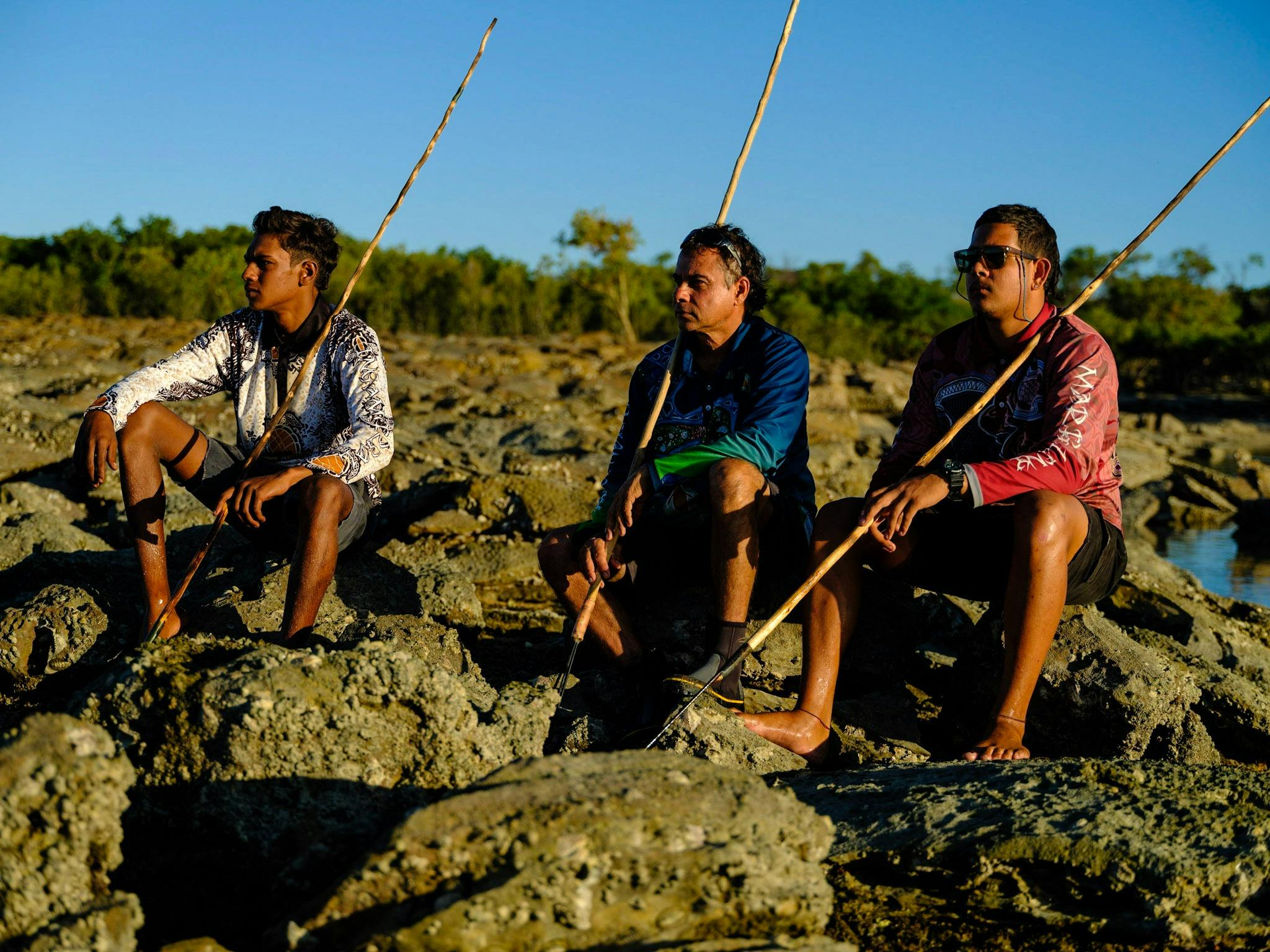 three people sitting on stones in mangrove wearing ardi'ol fishing shirts and holding spears.