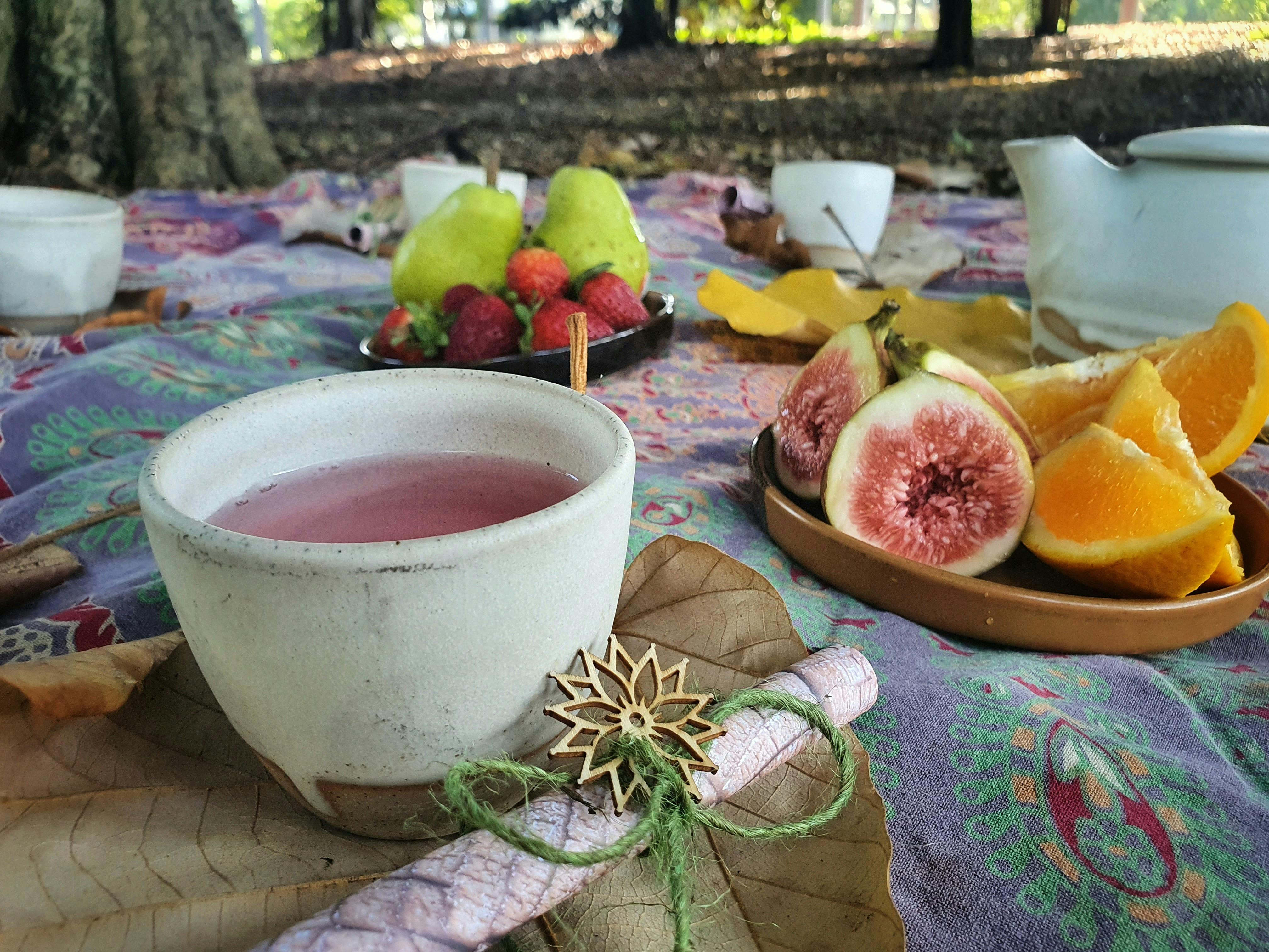 A cup of pink tea at a picnic, with figs and orange segments in the background.