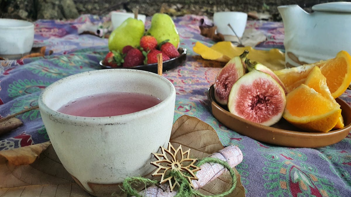 A cup of pink tea at a picnic, with figs and orange segments in the background.