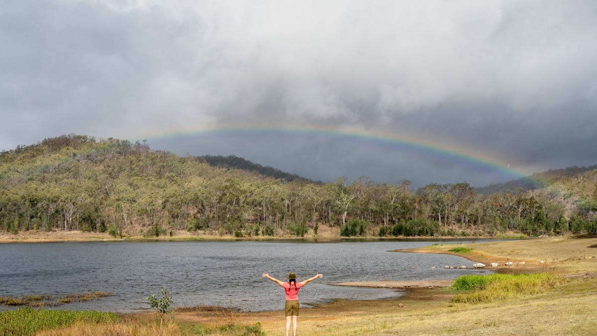 A girl holds her hands in the air as a rainbow is cast over Mount Morgan No. 7 Dam