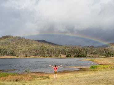 A girl holds her hands in the air as a rainbow is cast over Mount Morgan No. 7 Dam