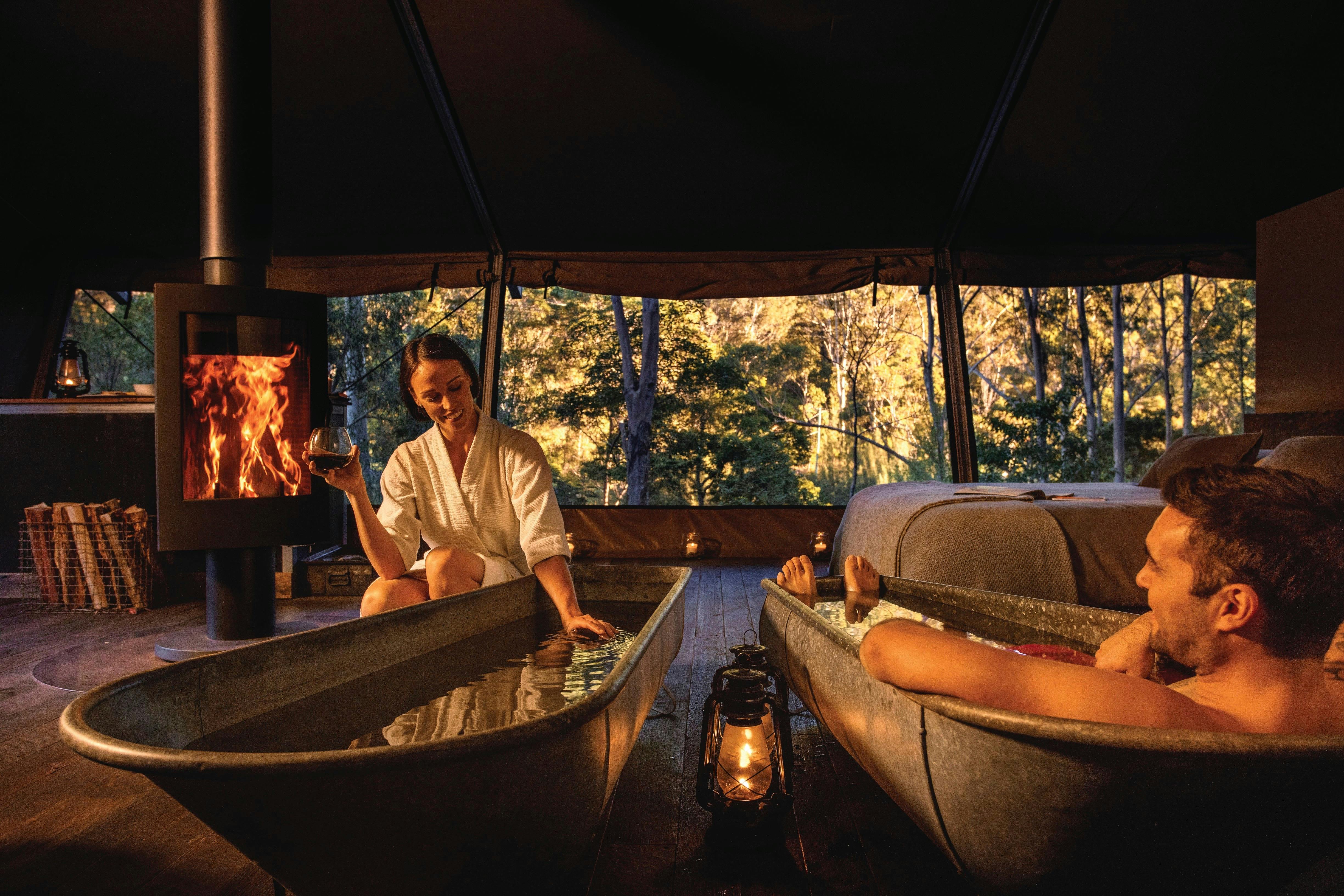 Couple enjoying a bath by the fire inside safari tent with view outside natural landscape.