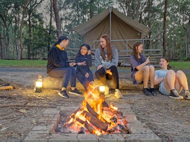 Five young girls sitting around a campfire at dusk, in front of a platform tent